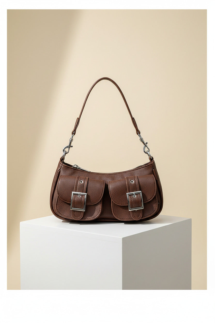 A person wearing a beige trench coat and blue striped shirt carries a brown leather shoulder bag with silver buckle details.